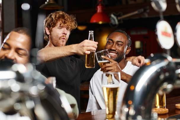 cheerful multiethnic friends hugging and toasting glasses of beer while spending time in bar - Photo, Image