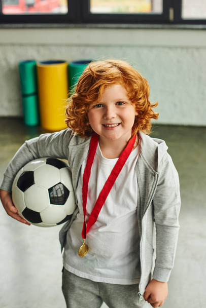tiro vertical de niño de pelo rojo bonito con medalla de oro sosteniendo pelota de fútbol y sonriendo alegremente - Foto, imagen