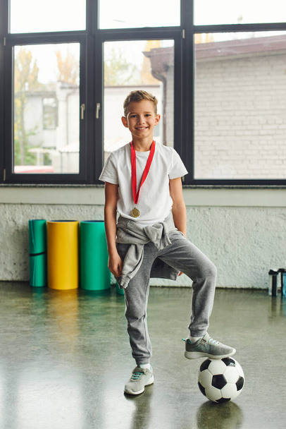 vertikální záběr roztomilého chlapce ve sportovním oblečení se zlatou medailí a fotbalovým míčem s úsměvem na kameru, sport - Fotografie, Obrázek