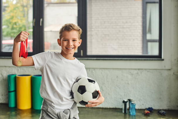 roztomilý veselý předpubertální chlapec ve sportovním oblečení drží zlatou medaili a fotbal, dětský sport - Fotografie, Obrázek