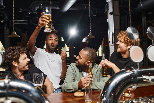 hombre afroamericano asombrado levantando un vaso de cerveza cerca de amigos alegres en el mostrador del bar, vida nocturna - Foto, Imagen