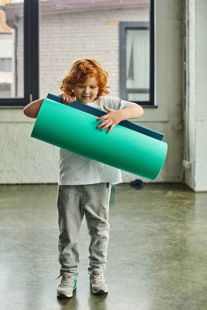vertical shot of cute boy with red hair in sportswear unfolding his karemat, child sport - Photo, Image