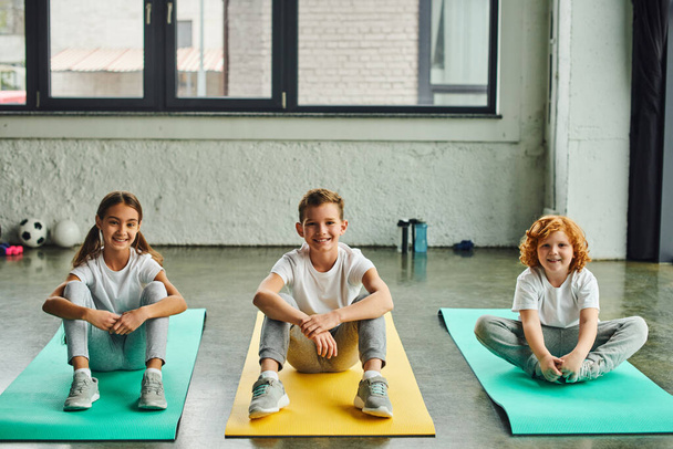 drei fröhliche vorpubertäre Jungen und Mädchen, die auf Fitnessmatten sitzen und in die Kamera lächeln, Sport treiben - Foto, Bild
