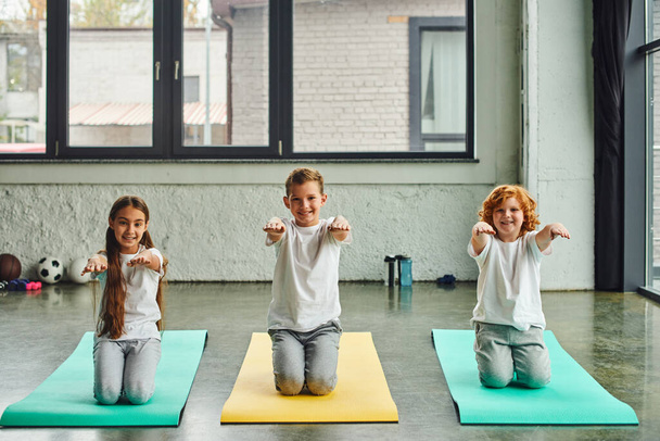 little joyful children sitting on fitness mats and stretching arms, smiling at camera, child sport - Photo, Image