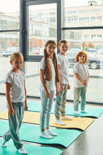 tiro verticale di bambini allegri in piedi su tappetini fitness e sorridente alla macchina fotografica, sport infantile - Foto, immagini