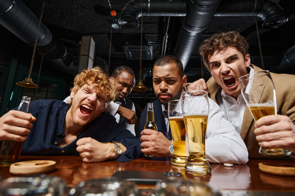 emotionele multiculturele collega 's in formele kleding die bier vasthouden en schreeuwen in de bar, mannen na het werk - Foto, afbeelding