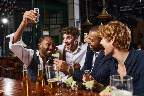 glücklich afrikanisch-amerikanischer Mann hebt Schuss von Tequila in der Nähe aufgeregter Freunde während Junggesellenabschied in Bar - Foto, Bild