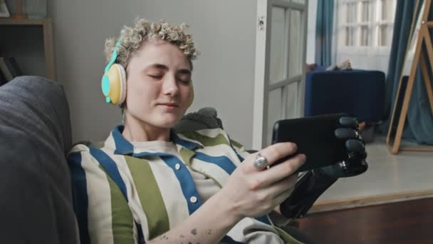 Stijlvol gen Z meisje met kort haar dragen arm prothese ontspannen in de woonkamer luisteren naar muziek in koptelefoon en surfen op internet op de smartphone - Video