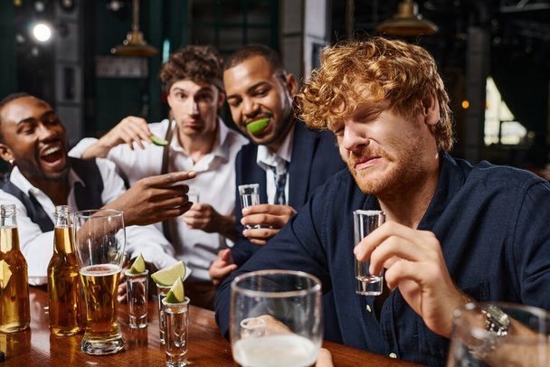 focus op roodharige man grimacing terwijl het drinken van tequila schot in de buurt van interraciale vrienden na het werk - Foto, afbeelding