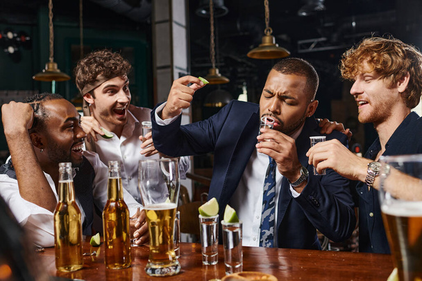 feliz interracial los hombres mirando africano americano amigo bebiendo tequila disparo y celebración de cal - Foto, Imagen