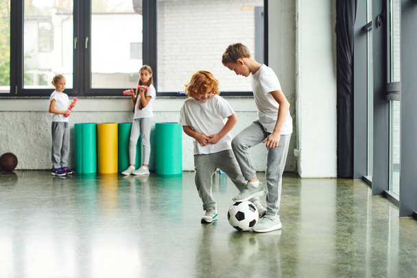 バックグラウンドでダンベルを保持する少女とサッカーをする陽気な小さな少年,子供のスポーツ - 写真・画像