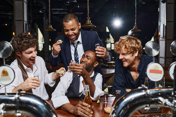 grupo de cuatro amigos multiétnicos felices y borrachos en traje formal beber tequila disparos en el bar - Foto, Imagen