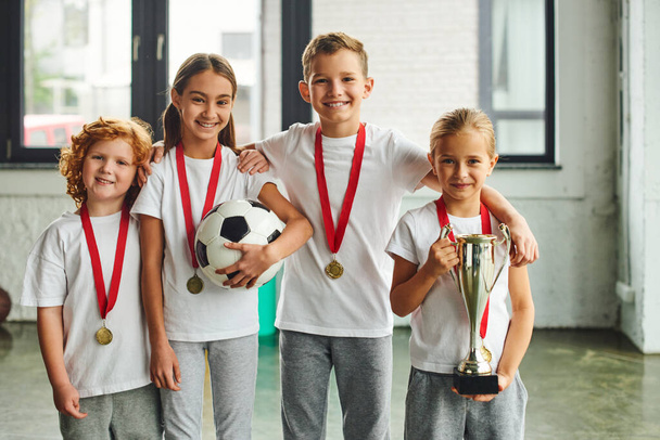 vidám kisfiúk és lányok kitüntetésekkel mosolyognak a kamera előtt, trófeát és focit tartanak a kezükben, sport - Fotó, kép