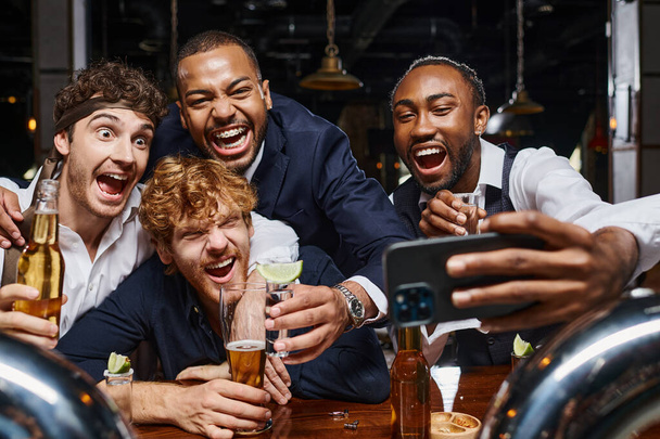 excited multiethnic colleagues in formal wear taking selfie on smartphone in bar after work - Photo, Image