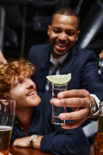 focus on tequila shot with sliced lime on top, african american man holding drink near friend in bar - Фото, изображение