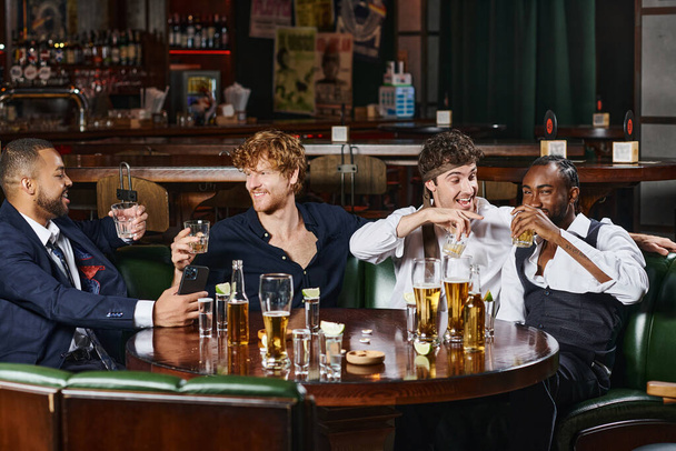 vier fröhliche multikulturelle Freunde trinken Whiskey in der Nähe von Tequila-Shots und Bier auf dem Tisch in der Bar - Foto, Bild