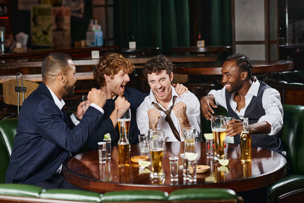 four happy and drunk interracial colleagues having fun after work in bar, alcohol drinks on table - Photo, Image