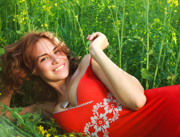 Smiling sexy young woman lying in the grass in a red dress - Foto, Imagen