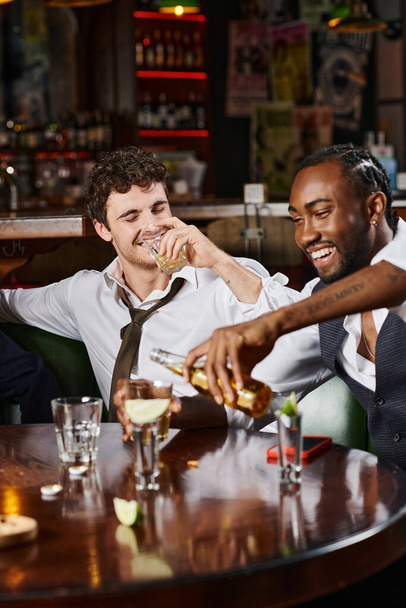 szczęśliwy pijany człowiek pije whisky w pobliżu wesoły afrykański przyjaciel wlewa piwo do szkła - Zdjęcie, obraz