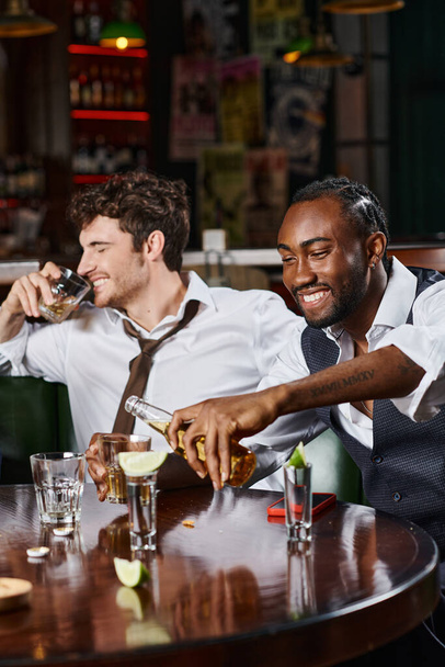 веселий афроамериканський чоловік виливає пиво в склянку біля друга, посміхаючись і п'ючи віскі - Фото, зображення