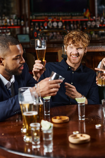 バーで仕事をした後,飲み物をしながら友人を指す陽気な赤毛の男,異人種間の友人 - 写真・画像