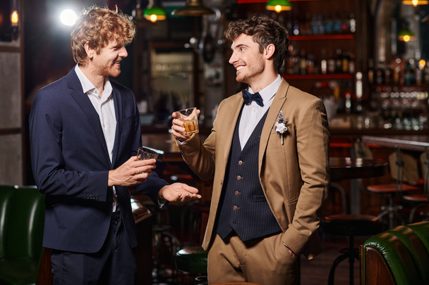 marié heureux en costume avec boutonnière et témoin tenant des verres de whisky pendant la fête de célibataire - Photo, image