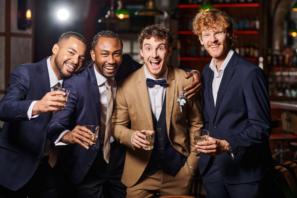 happy groom standing with interracial friends and holding glasses of whiskey during bachelor party - Photo, Image