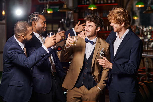 happy friends in formal wear congratulating groom in bar, interracial men holding glasses of whiskey - Photo, Image