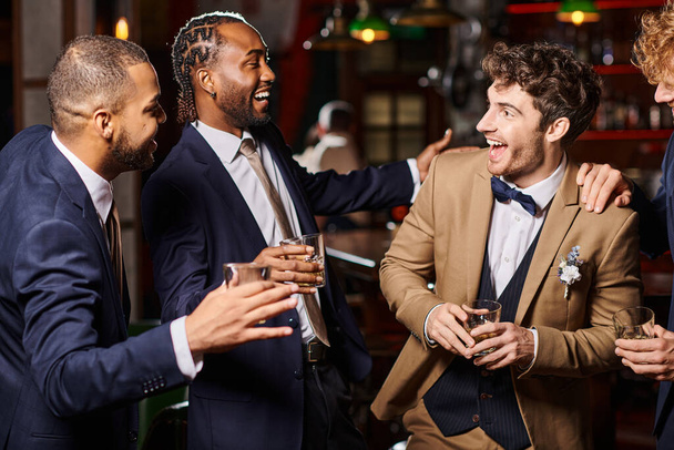 glückliche interrassische Freunde in formeller Kleidung gratulieren dem Bräutigam in der Bar, Männer halten Gläser Whiskey in der Hand - Foto, Bild
