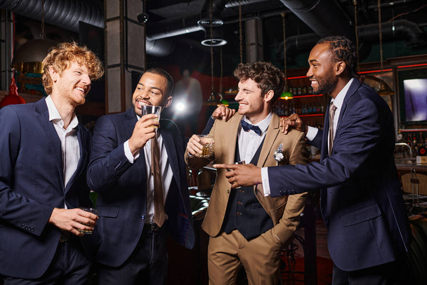 happy interracial best men in formal wear congratulating friend in bar, men with glasses of whiskey - Photo, Image