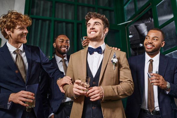 Junggesellenabschied, fröhliche gemischtrassige Männer gratulieren Freund in Bar, Trauzeugen und Bräutigam in Anzügen - Foto, Bild