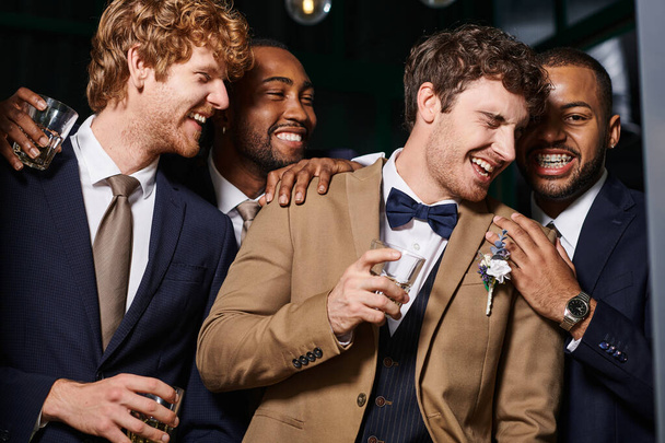 rozlučka se svobodou, nadšení multietničtí nejlepší muži a ženich smějící se v baru, držící skleničky whisky - Fotografie, Obrázek