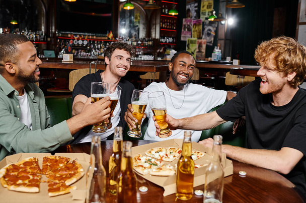 noc, szczęśliwy międzyrasowy mężczyźni brzęczenie szklanki piwa w pobliżu pizzy w barze, męska przyjaźń - Zdjęcie, obraz