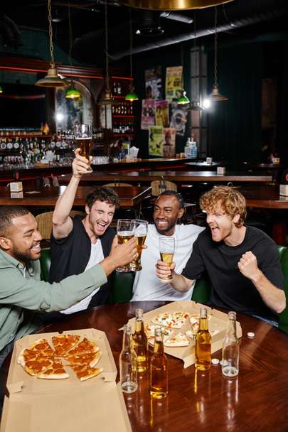 Nacht aus, aufgeregte interrassische Männer, die in der Nähe einer Pizza in einer Bar Biergläser klappern, Männerfreundschaft - Foto, Bild