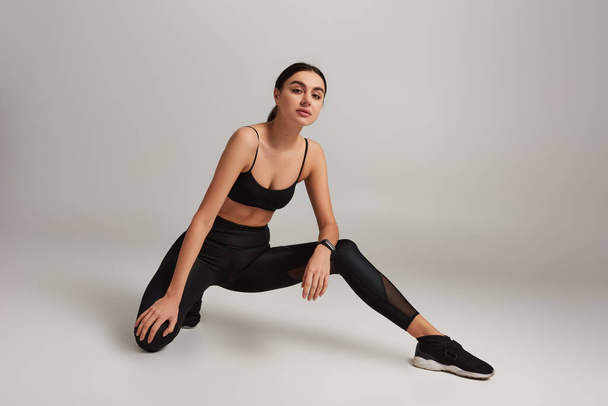 beautiful woman in black active wear with fitness tracker on wrist stretching leg on grey backdrop - Photo, Image