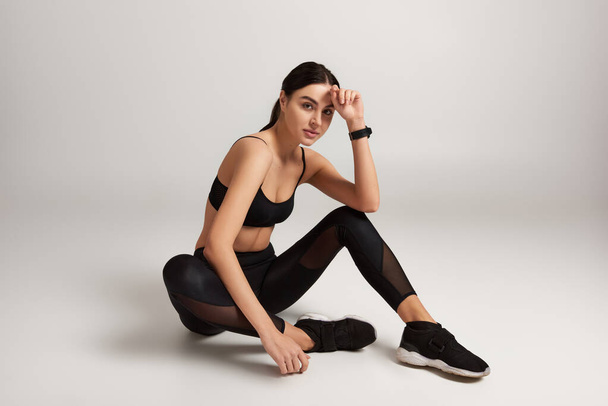 brunetka sportovkyně v černé aktivní oblečení s fitness tracker na zápěstí sedí na šedém pozadí - Fotografie, Obrázek