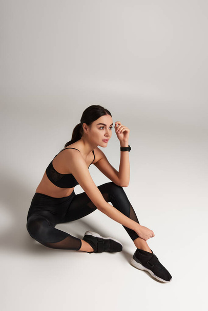 pensive sportswoman in black active wear with fitness tracker on wrist sitting on grey backdrop - Photo, Image