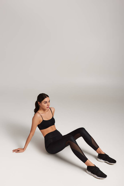 vermoeide sportvrouw in zwart actieve kleding met fitness tracker om de pols zittend op grijze achtergrond - Foto, afbeelding