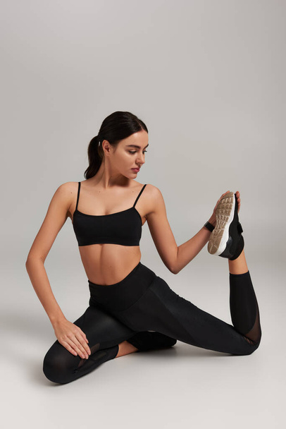 flexible and slim sportswoman in black leggings and crop top stretching legs on grey backdrop - Photo, Image