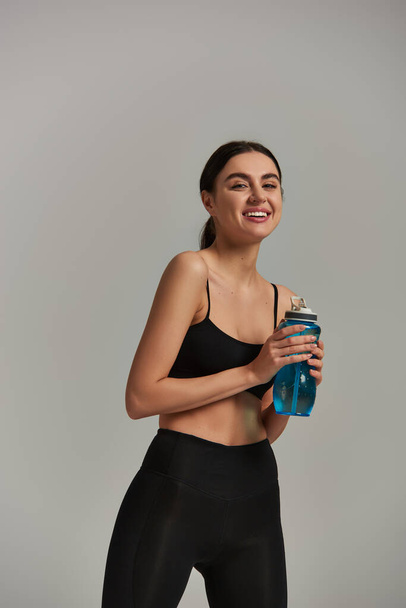 cheerful slim sportswoman in leggings and crop top holding bottle with water on grey background - Photo, Image