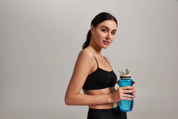 retrato de mujer en ropa deportiva sosteniendo botella con agua mientras está de pie sobre fondo gris - Foto, imagen