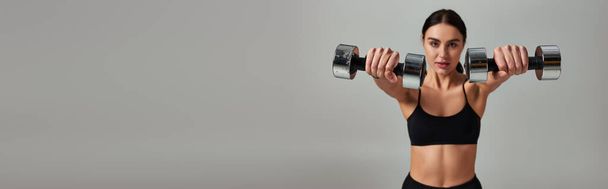 motivierte Sportlerin in schwarzem Aktiv-Outfit beim Training mit Kurzhanteln auf grauem Hintergrund, Banner - Foto, Bild