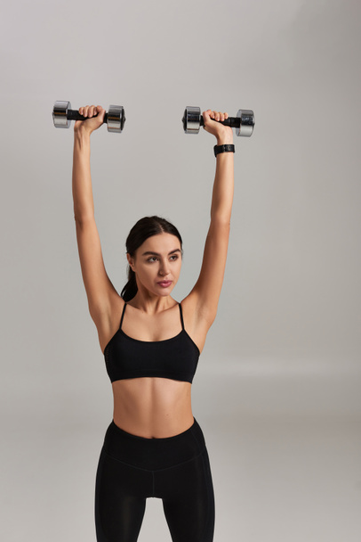 strong and sportive woman in black active wear lifting dumbbells above head on grey background - Photo, Image