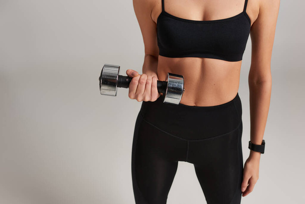 cropped slim and strong woman in active wear lifting dumbbell while working out on grey backdrop - Photo, Image