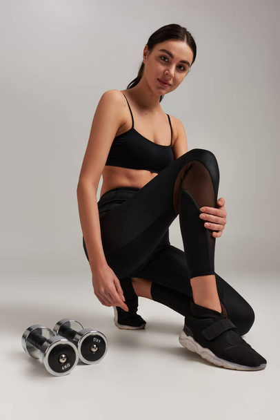 vékony és erős fiatal nő aktív kopás ül közelében súlyzók edzés után szürke háttér - Fotó, kép