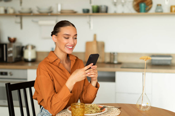 Radosna pani korzystająca ze smartfona, siedząca przy stole kuchennym i jedząca lunch, pisząca SMS-y na telefon komórkowy, surfująca po Internecie w domu, wolna przestrzeń - Zdjęcie, obraz