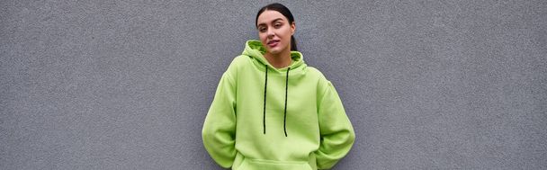 beautiful sportswoman in trendy lime color hoodie standing near grey concrete wall outdoors, banner - Photo, Image