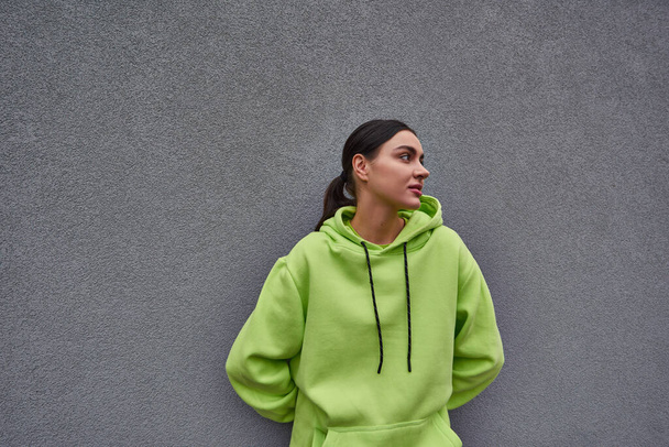 brunette young woman in trendy lime color hoodie standing near grey concrete wall outdoors - Photo, Image
