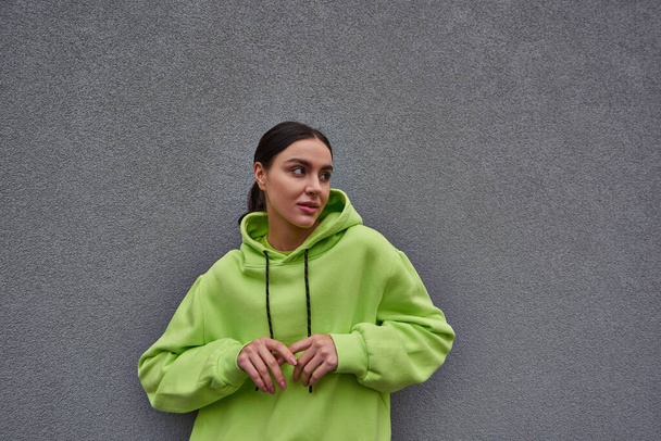 brunette young woman in trendy lime color hoodie looking away near grey concrete wall outdoors - Photo, Image