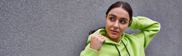 brunette young woman in trendy lime color hoodie posing while standing near concrete wall, banner - Photo, Image